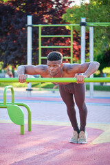 Young fit shirtless black man doing push-ups on a bar in a calisthenics park outdoors on sunny day....