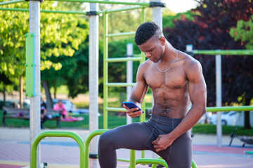 Portrait of a young fit shirtless black man using the smartphone in a calisthenics park outdoors....