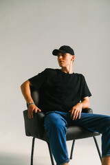 Portrait of handsome caucasian man fashion model in t shirt and cap sitting on chair, studio shot.