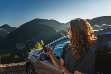 Young beautiful woman traveling by car in the mountains using smartphone at sunset, summer vacation...