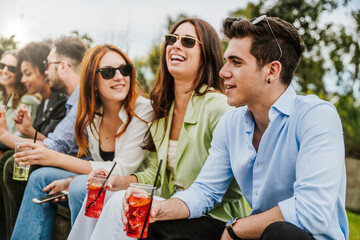 Cheerful best friends chatting and drinking fancy cocktails in plastic glasses sitting on a bench outdoors in the parkland - people unity and friendship lifestyle concept