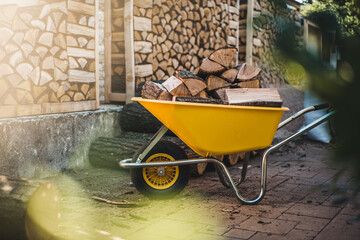 A pile of split firewood for heating a house in the yard, a yellow wheelbarrow with firewood in the foreground. - obrazy, fototapety, plakaty