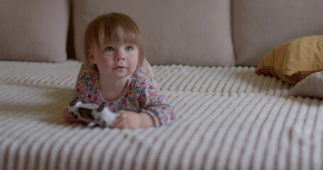 Fototapeta premium Cheerful little girl is lying on the sofa. She raises her head sharply, looks, smiles. Positive mood motivation. Relaxation of the child's psyche.