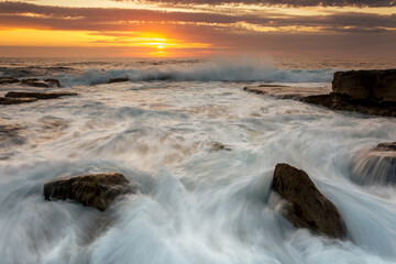 Incoming waves surge over rocks with a golden orange sunrise