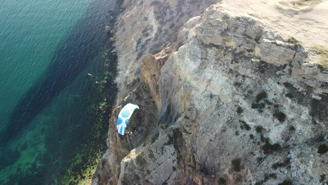 Aerial drone view of a man flying a white and blue paraglider over a hill and trees to the sea waves near the rocks. Active paraglider flight over the seascape with clear skies at suset. Extreme sport