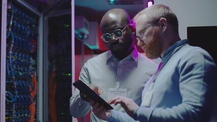 African American and Caucasian male colleagues discussing something on digital tablet while working together in server room of data center - Powered by Adobe