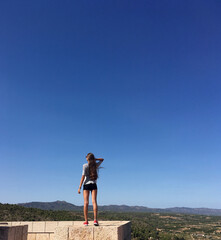Fototapeta premium A teenage girl with long hair stands on the stone wall of an old castle against a bright blue sky. Сoncept of freedom and inspiration. Travel content
