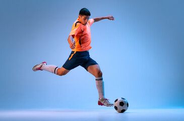 Fototapeta na wymiar Dynamic portrait of young man, professional football player in motion, training, dribbling ball isolated over blue studio background in neon light