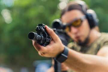 Man wearing camouflage t-shirt safety headphones and goggles aiming machine gun. Outdoor shooting...