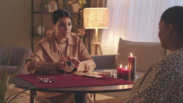 Waist up slowmo of Middle Eastern female fortune teller spinning crystal ball and smiling mystically while predicting future of African American woman sitting in front of her