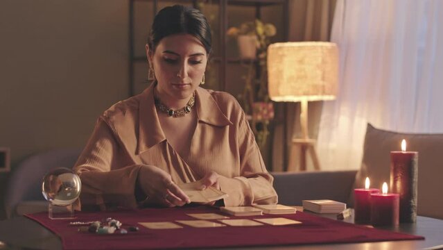 Waist up slowmo of young Middle Eastern fortune teller laying out tarot cards at table, sitting alone in dark room with burning wax candles