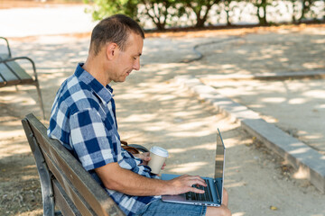 Man sitting on a bench drinking a drink from a paper cup, and working on a laptop 