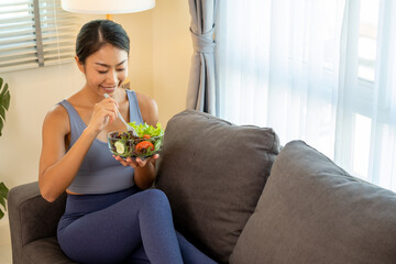 Happy women hold a salad bolw while sitting on a sofa in a living room.