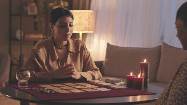Waist up slowmo of young Middle Eastern female fortune teller reading tarot cards to African American woman during seance