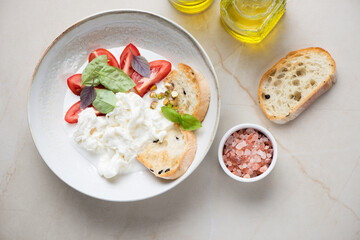 Plate with stracciatella or stretched curd cheese, ciabatta, tomato wedges and fresh basil, flat lay on a beige stone background, horizontal shot