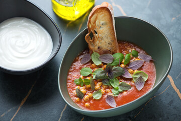 Tomato and chickpea soup served with fresh basil, ciabatta and yogurt, horizontal shot on a dark-olive marble background