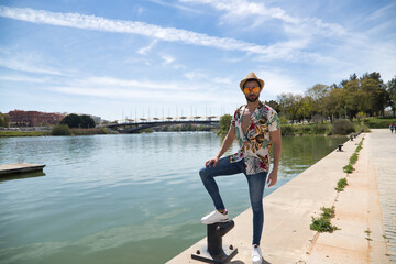 Young and handsome man, mirror sunglasses, beard, hat, Hawaiian shirt and jeans on a pier by the...