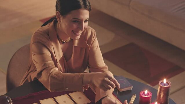 From above slowmo of young Middle Eastern fortune teller using tarot cards and pendulum while reading palm of African American woman sitting at table in front of her