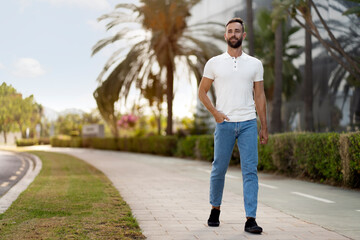 Fototapeta na wymiar Smiling handsome man wearing white t shirt, stylish jeans walking on the street, looking away. Copy space. Vacation, travel concept 