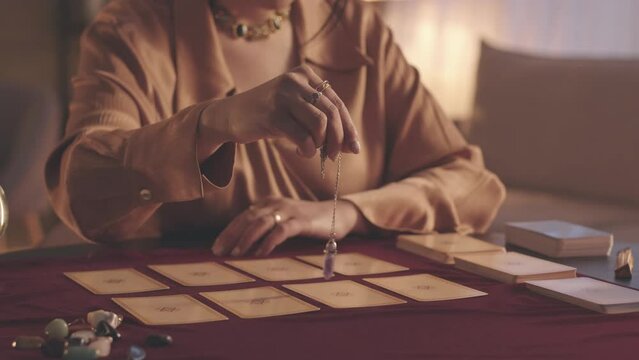 Cropped slowmo of unrecognizable fortune teller holding pendulum over tarot cards layout on table