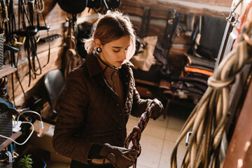 Young jockey woman preparing tack before horse riding in stable