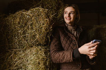 Young jockey woman using mobile phone while standing by hay in stable © Drobot Dean