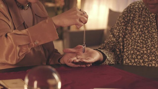 Cropped closeup slowmo of fortune teller with pendulum conducting palm reading of client during spiritual seance