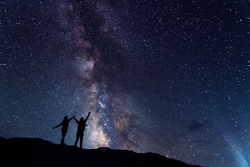 Two hiker silhouette stands on the hill and looking at the bright milky way galaxy in the starry...