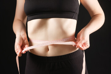 A woman measures the circumference of her abdomen on a black background. The concept of weight loss
