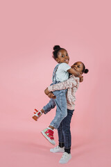 Funny two little African cute girls sisters, having fun together, posing to camera one girl rises other in her arms, on isolated pink color studio background.