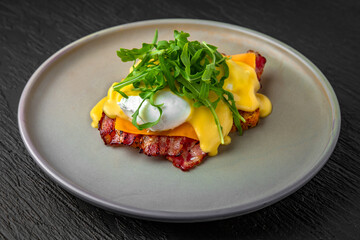 Healthy nutritious breakfast of toast, cheese, poached egg, bacon and greens in a ceramic plate on a dark textured background. Restaurant menu Isolated on black