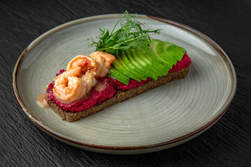 Healthy nutritious breakfast of bruschetta with beetroot paste, avocado and shrimp in a ceramic plate on a dark textured background. Restaurant menu Isolated on black