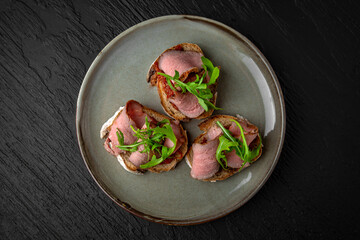 Healthy nutritious breakfast of bruschetta with pastrami beef ham and herbs in a ceramic plate on a dark textured background. Restaurant menu Isolated on black