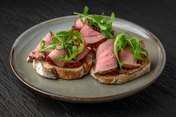 Healthy nutritious breakfast of bruschetta with pastrami beef ham and herbs in a ceramic plate on a dark textured background. Restaurant menu Isolated on black