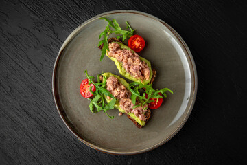 Cereal bread with tuna and avocado in a ceramic plate on a dark textured background. Restaurant menu Isolated on black