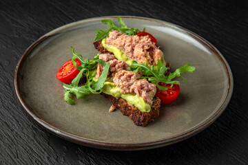 Cereal bread with tuna and avocado in a ceramic plate on a dark textured background. Restaurant menu Isolated on black