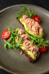 Cereal bread with tuna and avocado in a ceramic plate on a dark textured background. Restaurant menu Isolated on black