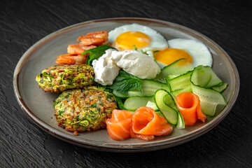 Hearty nutritious dietary breakfast of eggs, zucchini pancakes with salmon and shrimp in a ceramic plate on a dark textured background. Restaurant menu Isolated on black