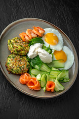 Hearty nutritious dietary breakfast of eggs, zucchini pancakes with salmon and shrimp in a ceramic plate on a dark textured background. Restaurant menu Isolated on black