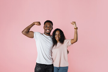 Young cheerful strong sporty fitness african couple two friends man woman in casual t-shirts showing biceps muscles on hand isolated on pastel pink color background studio portrait.