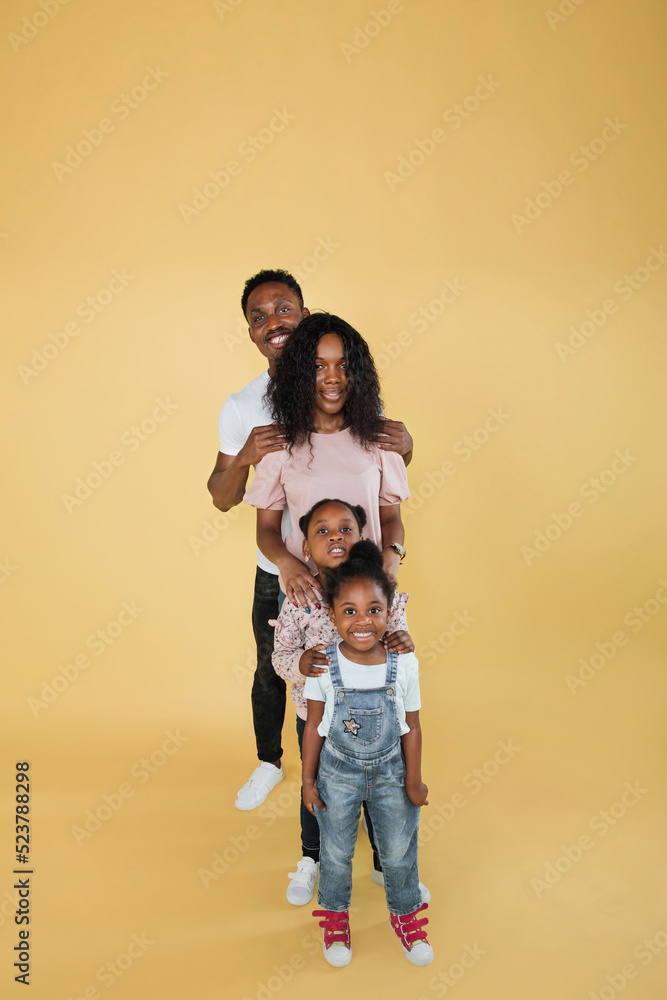 Wall mural Cheerful african american family of four, standing one behind other and smiling at camera on yellow background, banner, free space.