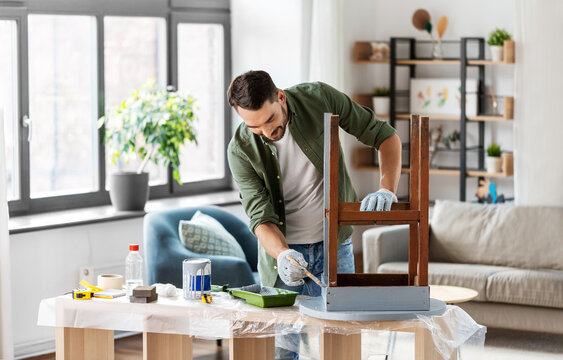 furniture renovation, diy and home improvement concept - happy smiling man in gloves with paint brush painting old wooden table in grey color