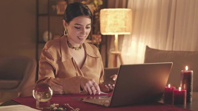 Waist up slowmo of young Middle Eastern fortune teller laying out tarot cards to client during online video seance on laptop