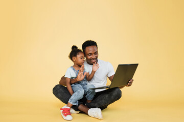 Horizontal shot of happy joyful African family, young father with cute little kid daughter, sitting on the floor and waving hands while watching a video lesson on laptop pc on yellow studio background
