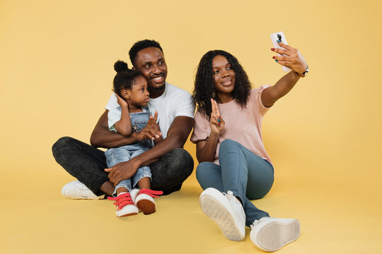 Happy Family, Technology, Communication Concept. Young African Parents Sitting On Floor With Their Daughter Showing Sign Of Peace And Embracing, Using Smartphone For Selfie On Yellow Background