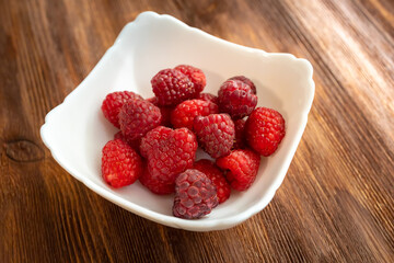 in a white plate, which stands on a wooden table, juicy fresh raspberries, on which the rays of the sun fall