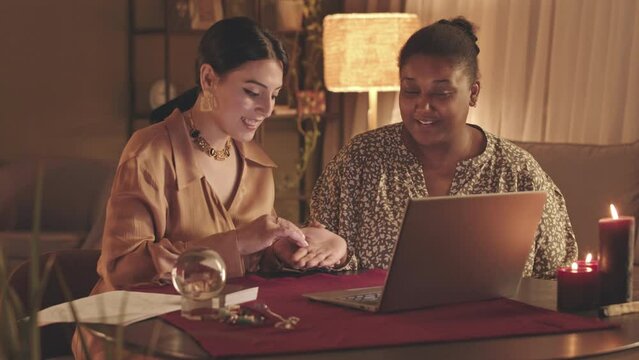 Waist up slowmo of fortune teller and client sitting in front of laptop while practicing chiromancy during live stream