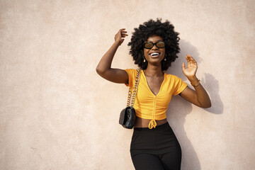 Happy African American young woman with afro hair wearing stylish sunglasses posing against beige...