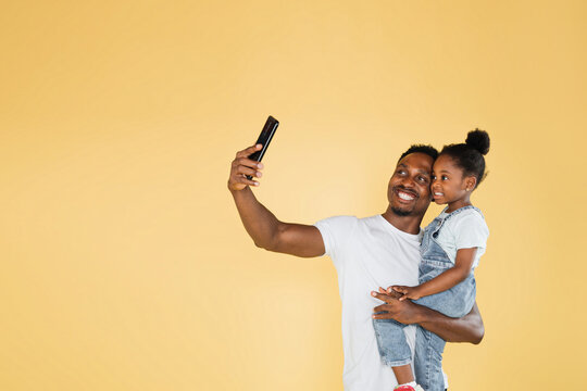 Happy African American family dad and daughter having fun and using mobile phone video call on yellow background. Self, isolated, social distancing, quarantine for coronavirus prevention.