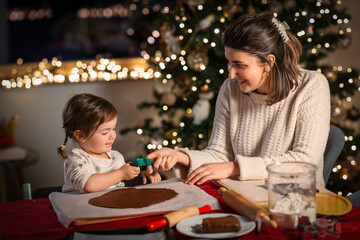 family, cooking and winter holidays concept - happy mother and baby daughter with mold making gingerbread cookies from dough at home on christmas